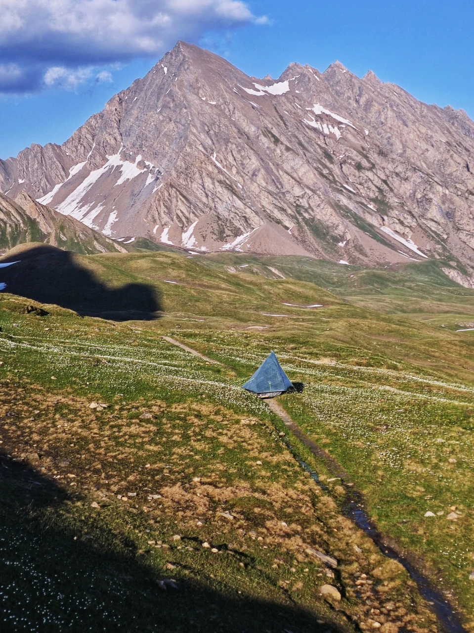A tent pitched on a TMB pasture in early June, tiny beneath the summit with snow still lingering: an atmosphere that no longer exists once high season is underway